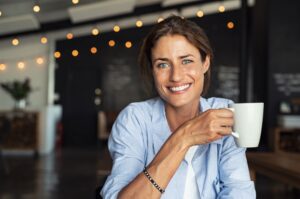 A woman drinking coffee and smiling at the camera, at a cafe with string lights in the background.